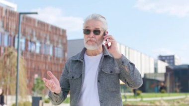 Gündelik kıyafetleri ve güneş gözlükleriyle cadde boyunca yürürken modern akıllı telefonuyla konuşan ciddi bir yaşlı insan. Kasaba atmosferindeki önemli meselelere odaklanan aktif sakallı adam..