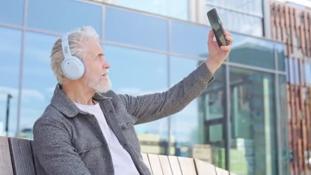 在现代城市公园里 人们一边享受着好天气 一边试图用新型智能手机自拍 高级男子在无线耳机外使用数字小玩意 — 图库视频影像