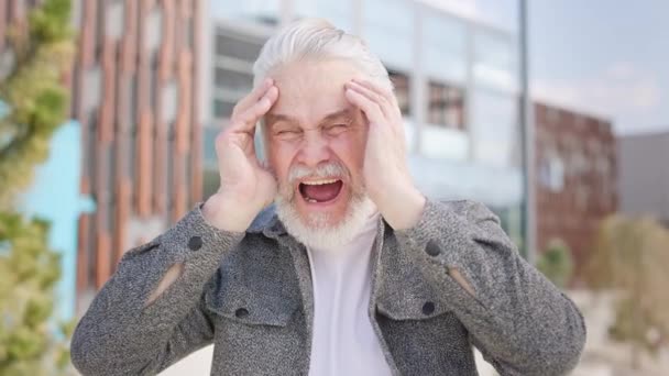 年长男性 灰胡子 双手抱头 站在城市公园里痛得大叫 穿着外套 衣着时髦的退休者站在外面头疼难忍的画像 — 图库视频影像