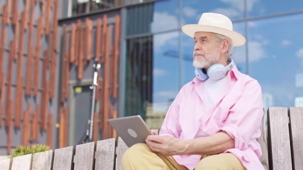 坐在木制长椅上 手上拿着数码平板电脑 头戴雅致的白帽 自信的老年男性侧视图 严肃的高加索人 用现代小玩意在新鲜空气上消磨时光 — 图库视频影像