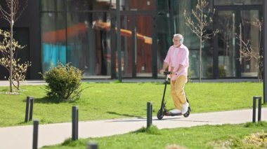 Modern şehir parkında elektrikli scooter kullanan yaşlı bir adamın yan görüntüsü. Gri saçlı erkek, günlük kıyafetler ve kablosuz kulaklıklar takıyor. Çevre taşımacılığını kullanarak doğayla ilgileniyor..