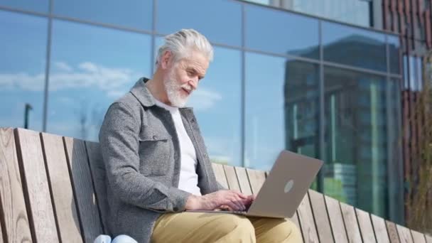 身穿休闲装的快乐老年高加索男性坐在木制长椅上 膝上型无线膝上型笔记本电脑 双手张开 在开阔的空间上显示出快乐 成绩概念和技术 — 图库视频影像