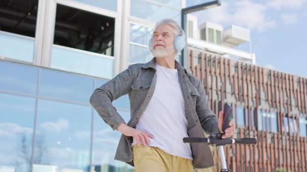 見栄えの良い引退した男性は Eスクーターが都市の背景に注意深く何かを見ているカジュアルな服で 成功した灰色の髪の男性携帯電話を保持し 思慮深く距離を調べる — ストック動画