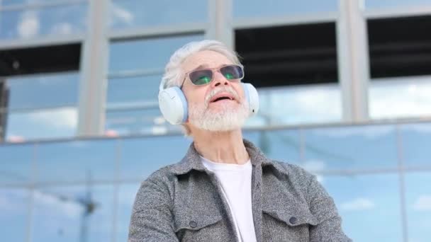天気の良い場所を賞賛する灰色のひげを持つ上級男性の肖像画 現代都市の雰囲気の中で素晴らしい時間を過ごすワイヤレスヘッドフォンとサングラスを備えたアクティブな退職者 — ストック動画