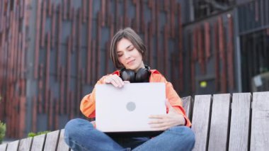 Modern bina arka planında otururken dizüstü bilgisayarı açıp kollarını ve kollarını açarak şık bir esmer. Uykulu bayan üniversitede online derslere hazırlanıyor. E-öğrenme kavramı.