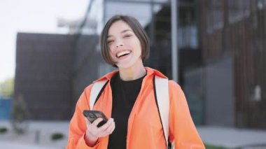 Çekici kadın portresi, şehir bölgesinde olumlu duyguları ifade ederken parmaklarıyla iyi bir hareket gösteriyor. Beyaz kadın, günlük kıyafetlerle akıllı telefon ekranına bakıyor ve tavsiye mektubu gösteriyor..