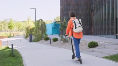 Modern bir kadının elektrikli scooterla modern şehir parkında seyahat edişinin arka görüntüsü. Çekici beyaz kadın şehir manzarasında taşınmak için çevre dostu ulaşım kullanıyor..
