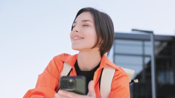 パーソナルスマートフォンを持っているカジュアルなオレンジのジャケットに身を包んだ美しい女性のクローズアップと晴れた通りに立っている間脇を見ます 屋外の現代ガジェットを使用してかなり慎重な女性 — ストック動画