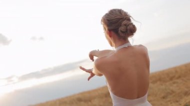 Beyaz, bol elbiseli zarif bir bayanın geniş bir alanda zarif bir şekilde hareket edişinin arka görüntüsü. Beyaz bir kadın yaz günbatımında kollarını açıyor ve arkasına yaslanıyor..