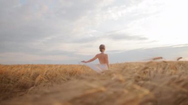 Uzun ve altın buğdaylı uçsuz bucaksız tarlaların arasında dans eden çekici beyaz kadın. Nazik, gevşek, beyaz elbiseli kadın açık alanda güzel bir şekilde hareket ediyor. Gün batımında bulutlu mavi gökyüzü.