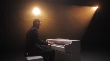 Kostümlü bir adamın sahne ışığında piyanonun başında oturması ve konser sırasında müzik çalması. Yetişkin erkek müzisyen sahnede performans sergiliyor ve klasik melodinin seslerinden tatmin oluyor..