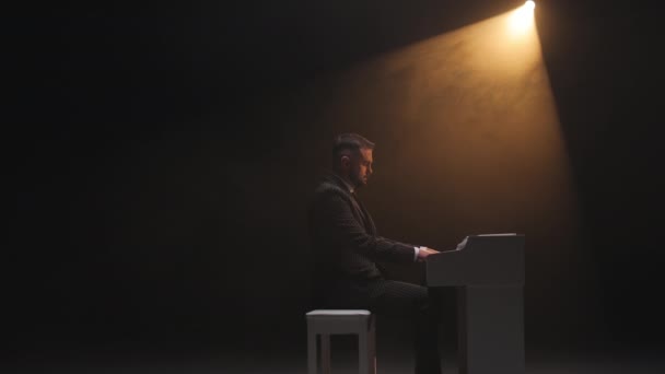 在音乐会节目中以新歌表演的著名男歌手的侧视图 男人坐在钢琴前 在大厅里弹奏着迷人的乐器曲调 还有对比灯光 — 图库视频影像