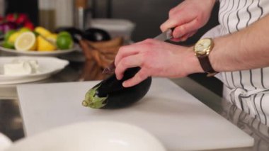 Şef çizgili önlük giymiş mutfak ortamında beyaz tezgahta patlıcan kesiyor. Bıçak tutan ve sebzeleri ikiye bölen bir erkek. Limonlu ve limonlu kasenin arka planı.