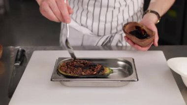 Yetkin şef çizgili önlük giyer ve diğer elinde küçük bir kâse tutarken yemekleri kaşıkla süsleyip baharat katar. Erkek, içeride ızgara patlıcan hazırlıyor..