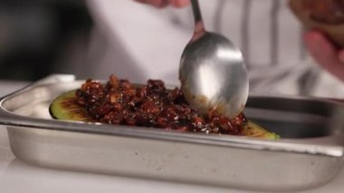 Mutfakta kaşıkla ızgara patlıcanın üstüne sos koyan usta bir şef. Önlük giyen çalışkan aşçılık uzmanı metal tepside sebzelerle çalışıyor..
