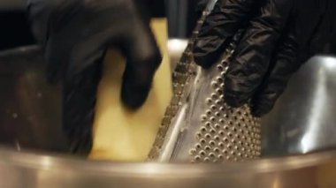 Profesyonel şefin siyah eldivenler giyip metal rende peynir rendeleyerek paslanmaz çelik kaseye dönüştürdüğü yakın plan. Bileşen parçalarla rendeleme eylemine odaklanmış.