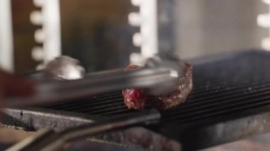 Şef, ızgara ızgara izleriyle et parçasını kızgın ızgarada pişerken metal maşayla çeviriyor. Malzemelerin işleme konsepti, hazırlık ve restoran.