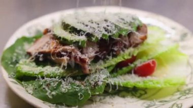 Beyaz tabakta rendelenmiş peynirle kaplı yeşil marul yapraklı taze salata ve dilimlenmiş domatesler. Çiçeklerle servis edilen sebzelerin altında ızgara et..