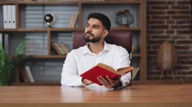 Beyaz tişört giymiş neşeli iş adamı elinde kırmızı kitapla yan tarafa bakıyor. Düşünceli Hintli erkek dekoratif eşyalarla rafların arkasında metni düşünüyor..