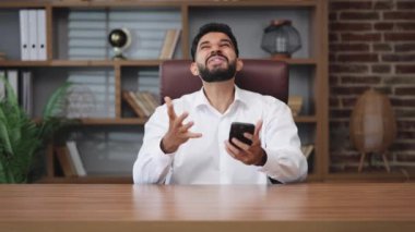 Heyecanlı Hintli erkek yumruklarını sıkarak ve olumlu jestler yaparak ofis ortamındaki kişisel akıllı telefonlara bakıyor. Enerjik bir adam kendini iyi hissediyor..