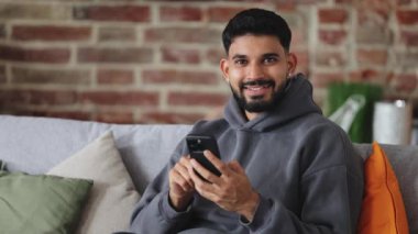 Kameraya dostça gülümseyerek bakarken akıllı telefonu kollarında tutan mutlu Arap erkek portresi. Rahat giysiler içinde iyimser bir insan evinde modern cihaz kullanıcısı deneyiminin tadını çıkarıyor.