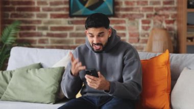 Sabırsız Arap adam saldırganlıkla bağırıyor ve akıllı telefonu oturma odasındaki kanepeye atıyor. Stresli erkek, aletlere yumruk atmaktan kaçındıktan sonra elleri ile yüzünü kapatıyor..