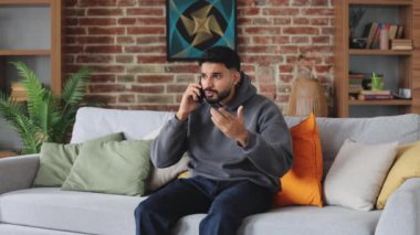 Gündelik giysiler içinde endiseli bir erkek, önemli bilgileri modern akıllı telefondan aktarıyor. Gergin adam evde koluyla el kol hareketi yaparken elektronik cihazı kulağından tutuyor..