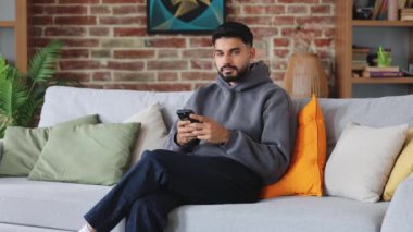 Endişeli yetişkinler modern telefon kullanmaktan ve kameraya dikkatlice bakmaktan çekiniyorlar. Günlük giysiler içinde çekici bir adam oturma odasında sosyal medya yayınına ara veriyor..