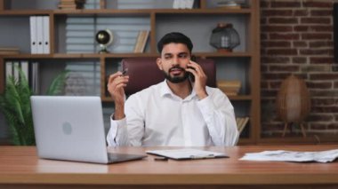Odaklanmış Hindu erkek portresi açık kablosuz dizüstü bilgisayarın yanındaki belgelere bir şeyler yazarken modern akıllı telefon konuşması yapıyor. Kapalı alanda hoş bir ayrılık yaşayan olumlu bir adam..