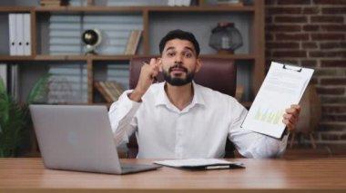 Ofis ortamında kişisel işyerinde otururken belgelere kalem tutan duygusal Hindu erkek işçi. Çalışmak için peronal dizüstü bilgisayar ve ekipman kullanan enerjik erkek.