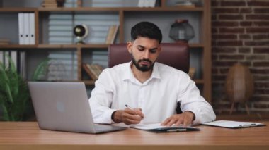 Ofisteki modern dizüstü bilgisayarın klavyesinde yazı yazan çalışkan Hintli bir erkeğin ön görüşü. Beyaz gömlekli ciddi bir adam evdeki belgelere not alıyor..