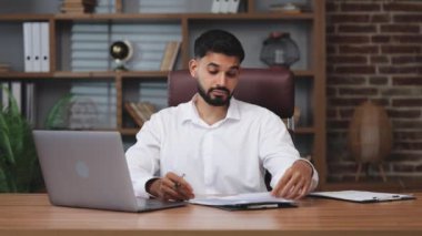 Şaşkın erkek, iş saatinin sonunda kablosuz bilgisayarı kapatıyor ve içerideki ahşap masaya uzanıyor. Uykulu Hintli adam ofisteki kişisel işyerinde dinleniyor. Rahatlama kavramı.