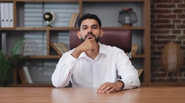 Küçük Hintli girişimci kişisel kabinede doğrudan kameraya bakarken gülümseyerek çenesine dokunuyor. Evin içinde gömlek giyerken resmi giyinmiş kendinden emin bir erkek..