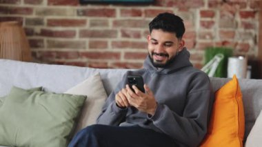 Gri kapüşonlu pozitif adam evde dinleniyor ve kablosuz internet bağlantısı olan modern bir cihaz kullanıyor. Tuğla duvarın arkasındaki akıllı telefon ekranına ilgi duyan sakallı bir adam..