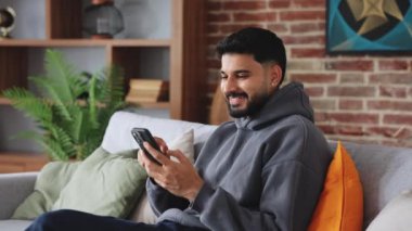 Dairesinde yastıklı kanepeyle yeni cep telefonu kullanırken gündelik giysiler giyen tatmin olmuş bir adam. Kulak delen mutlu kişi, arkadaşlık mesajından hoş haberler okuyor ve internet sitesine bağlanıyor..