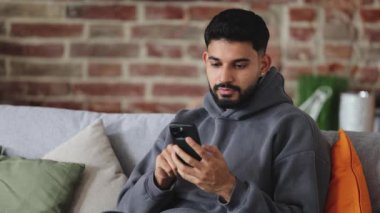 Sade tişörtlü Hintli adam siyah çantalı, yüzünde sakince bir ifade olan akıllı telefona bakıyor. Yastıklı kanepede otururken, blogcudan gönderi arayan odaklanmış insanlar.