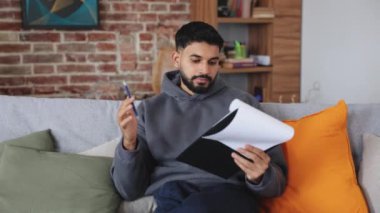 Koyu renk kapüşonlu ciddi bir adam kanepede oturuyor ve tablete bir sürü kağıtla bir şeyler yazıyor. Özenli kişi anket veya iş başvurusu doldurup soruya işaretleme yanıtı verir.