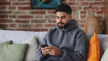 Koyu gri kapüşonlu bir erkek akıllı telefon ekranına bakıyor ve konsantre yüzüyle kameraya bakıyor. Kulak delici, yeni aletler test eden ve detaylara dikkat eden ciddi bir adam..