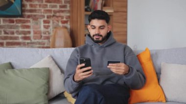 İçerideki rahat mobilyaların üzerinde otururken kredi kartı ve cep telefonuna bakan konsantre bir erkek. Çevrimiçi bankacılık hizmetinin genç müşterisi hesap kaydı için ayrıntıları giriyor.