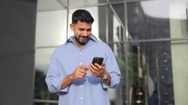 Çekici sakallı adam, günlük kıyafetlerle internette cep telefonuyla geziniyor, şehirde duruyor. Akıllı telefon kullanan, arkadaşlarıyla sohbet eden, gülümseyen beyaz bir adamın alçak açılı görüntüsü. Teknoloji kavramı.