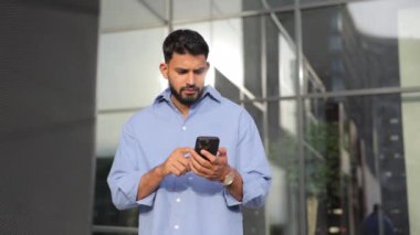 Resmi giysiler içinde sakallı yakışıklı bir adam cep telefonuyla yazı yazıyor, şehir caddesinde duruyor. Düşük açılı, koyu saçlı, yaz günü akıllı telefon kullanan adam. Teknoloji kavramı, bağlantı.