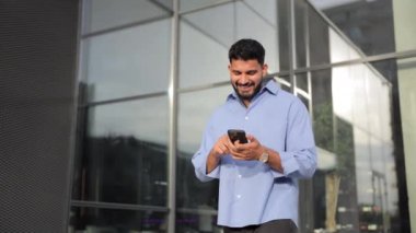 Şehirdeki iş yerinin önünde cep telefonu kullanan genç bir girişimci. Düşük açılı bir bakış açısıyla gülümseyen beyaz adam telefonda konuşurken, yürürken. İş, yaşam tarzı konsepti.