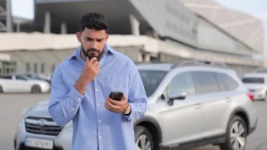 Kafası karışmış sakallı bir adam dışarıda telefon ekranına bakarken çenesini ovalıyor. Çekici erkek sürücünün gri arabaya karşı cep telefonu tutarken düşündüğünü, fikir sahibi olduğunu gösteriyor. İlham konsepti.