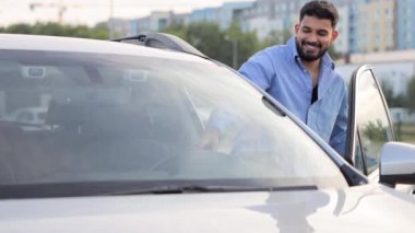 Gündelik kıyafetler giyen yakışıklı bir adam modern gri bir arabaya biniyor. Güvenlik kemeri kullanarak aracın kapısını açan mutlu, koyu renk saçlı erkek araba sahibinin yan görüntüsü. Araba kullanma konsepti.