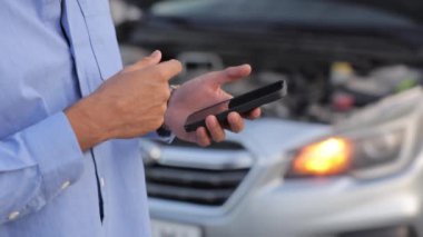 Mavi tişörtlü, cep telefonlu, servis numarasını arayan isimsiz bir erkek sürücü. Arka planda kırık bir arabayla elinde akıllı telefon tutan endişeli bir adamın görüntüsü. Araba bakımı kavramı.