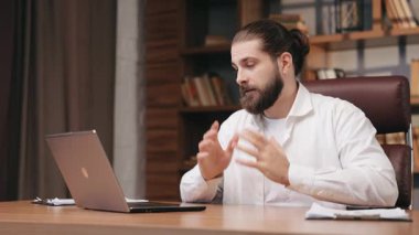 Yakışıklı sakallı iş adamı merkez ofisteki video konuşmalarıyla konuşuyor. Yöneticinin uzun saçlarıyla çöreğe bağlanmış, internette sohbet ederken, grafikleri ve grafikleri hareket halinde gösterirken yan görüntüsü. İş konsepti.