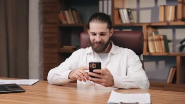 Uzun saçlı sakallı adam, ahşap masada oturuyor, kapalı alanda akıllı telefon kullanıyor. Ofiste dinlenirken telefon ekranına bakan gülümseyen orta yaşlı hippi, arka planda kitaplıklar. İş kavramı.