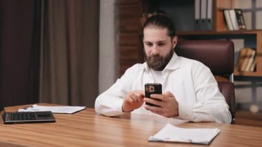 Sandalyede oturan, uzaktan çalışan ciddi bir adam. Gündelik giyinen, akıllı telefona kayan, ekranda hareket halindeyken sakala dokunan şaşkın erkek serbest yazar portresi. Duygular, iş kavramı.