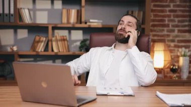 Gülümseyen, sakallı girişimci deri koltukta oturuyor ve uzaktan çalışıyor. Beyaz gömlekli iş adamının portresi, evde akıllı telefonla konuşuyor, raflara karşı, hareket halinde. Uzak çalışma konsepti.