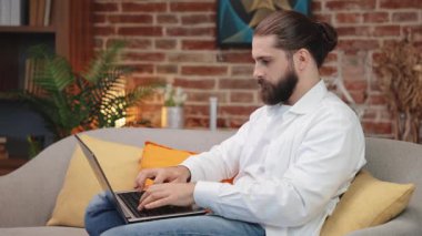 Laptop klavyesinde sıradan kıyafetlerle daktilo eden, koltukta oturan, çalışan bir adam. Uzun saçlı, ellerini ovuşturan, düşünen, serbest bir proje üzerinde çalışan adamın yan görünüşü. Uzak çalışma konsepti.